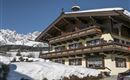 A traditional alpine house covered in snow during winter. In the background, majestic mountains can be seen under a clear sky.