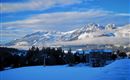 A winter landscape with snow-covered mountains and a clear blue sky. In the foreground is a snow-covered area, while clouds hang over the mountains.