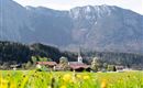 A picturesque landscape with blooming meadows and a small village in the foreground. In the background, majestic mountains rise under a clear sky.