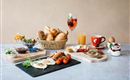 Ein reichhaltiges Frühstücksbuffet mit verschiedenen Brötchen, Eiern, Tomaten und einer Auswahl an Getränken. Dazu frisches Obst und süße Aufstriche.