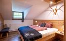 Een gezellige slaapkamer met houten panelen en een groot bed. Aan de wand hangt een geweer, en het raam laat veel daglicht binnen.