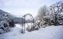 Eine schneebedeckte Landschaft mit einem Gartenbogen und verschneiten Pflanzen. Der Himmel ist grau und die Berge sind in der Ferne sichtbar.