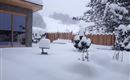 A snow-covered landscape with a wooden cabin and snow-covered trees. The snow lies thick on the ground and covers the garden furniture.