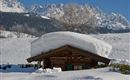 Eine winterliche Landschaft mit einem kleinen Holzhaus, das unter einer dicken Schneeschicht verborgen ist. Im Hintergrund sind hohe Berge und ein klarer blauer Himmel sichtbar.