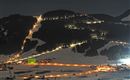 Een besneeuwd landschap bij nacht met verlichte skipistes en kleine huisjes. De lucht is donker en de lichten van het dorp verlichten de scène.