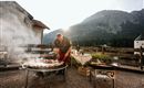 Ein Grillmeister bereitet verschiedene Speisen im Freien zu, umgeben von einer malerischen Berglandschaft. Der Rauch steigt in die Luft, während die Sonne im Hintergrund scheint.