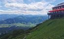Eine Berglandschaft mit einem Restaurant, das über eine Terrasse mit roten Schirmen verfügt. Die grünen Wiesen und das weitläufige Tal bieten einen schönen Blick auf die umliegenden Berge.
