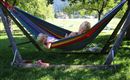Twee kinderen liggen in een kleurrijke hangmat onder een boom. De omgeving is groen en zonnig, met uitzicht op een weide op de achtergrond.