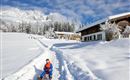 Ein Junge sitzt auf einem Schlitten in einer schneebedeckten Landschaft. Im Hintergrund sind schneebedeckte Bäume und gemütliche Häuser zu sehen.