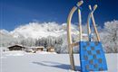 Ein Schlitten steht im frischen Schnee vor einem malerischen Bergpanorama. Im Hintergrund sind einige Hütten und schneebedeckte Bäume zu sehen.