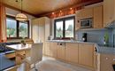 Eine moderne Küche mit Holzdecke und großen Fenstern. Der Essbereich hat einen Tisch und Stühle, sowie eine Küchenzeile mit verschiedenen Geräten.