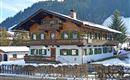 A traditional alpine house with a wooden balcony and green shutters. The garden is surrounded by snow-covered areas and wooded hills can be seen in the background.