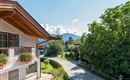 A charming landscape with a modern house and a beautiful garden. In the background, mountains and a clear blue sky can be seen.
