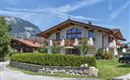 A beautiful, modern house with a well-maintained garden. In the background, mountains can be seen under a clear sky.