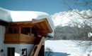 Ein schönes Holzhaus im Schnee mit einem geneigten Dach. Im Hintergrund sind beeindruckende Berge und ein blauer Himmel zu sehen.