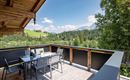 A cozy terrace with a table and chairs, surrounded by green forests and mountains. The sky is blue with white clouds.
