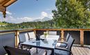 A cozy balcony with a table and chairs, surrounded by green mountains and trees. The sky is blue with some clouds.