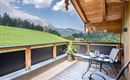 A cozy balcony with a table and chairs. In the background, green hills and mountains can be seen under a blue sky.
