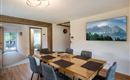 A modern dining room with a large wooden table and six chairs. A picture of mountain landscapes hangs on the wall, while large windows offer a view outside.