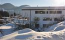 A snowy landscape with a modern building in the foreground. In the background, gentle hills and a clear sky can be seen.