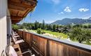 A balcony with a wooden railing and cozy chairs. In the background, there is a green meadow and mountains under a blue sky.