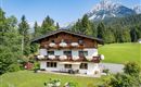 A charming alpine house with flower boxes and a large garden. In the background, majestic mountains and a clear sky can be seen.