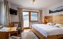 A modern bedroom with a large bed and wooden furniture. There is a table with chairs and large windows that offer a view of the mountains.