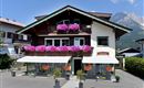A traditional Alpine house with colorful flowers on the balconies. In the background, mountains and a clear sky are visible.