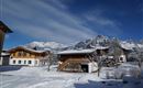 Eine winterliche Landschaft mit schneebedeckten Häusern und Bergen im Hintergrund. Der Himmel ist blau mit einigen Wolken.