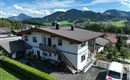 Ein modernes Haus umgeben von grünen Hügeln und Bergen. Der Himmel ist blau mit ein paar Wolken.<br/>Das Haus der Wohnung und im Hintergrund der Blick nach Ellmau.