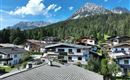 Eine malerische Alpenlandschaft mit traditionellen Häusern und dichten Wäldern. Im Hintergrund erheben sich majestätische Berg den Wilden Kaiser unter einem klaren blauen Himmel.