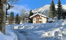 A picturesque winter landscape with snow-covered trees and a cozy house. The sky is clear and blue, in the background snow-covered mountains rise up.