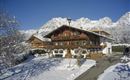 Ein traditionelles, holzverziertes Bauernhaus in einer schneebedeckten Landschaft. Im Hintergrund erheben sich majestätische Berge unter klarem blauen Himmel.