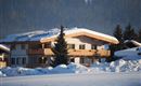 A cozy house in the snow with a large balcony. In the background, snow-covered trees and mountains can be seen.