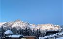 A snowy landscape with mountains in the background and snow-covered huts in the foreground. The sky is clear and blue.