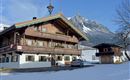 Ein traditionelles Holzhaus im Winter mit Schnee bedeckt. Im Hintergrund sind majestätische Berge und ein blauer Himmel zu sehen.