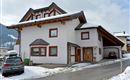 A modern house in the mountains, surrounded by snow. The facade is white with wooden details and large windows.