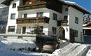A modern house in the snow with wooden-clad balconies. The parking lot is covered with snow and the surroundings show wintery mountains.