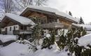 A rustic wooden house in the snow, surrounded by tall coniferous trees. The sky is clear and a peaceful winter landscape can be seen.