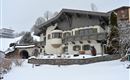A charming alpine house in the snow, surrounded by snow-covered mountains. The architecture is traditional with a charming bay window and wooden embellishments.