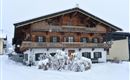 A traditional Alpine house with a wooden structure, surrounded by snowy terrain. The sky is gray and there are snowdrifts in front of the building.