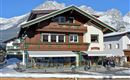 A traditional chalet with wooden cladding and balconies. In the background, snow-covered mountains and a clear blue sky are visible.