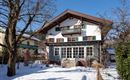 A charming alpine-style house with black-glossy windows. Surrounded by snow and trees, under a clear blue sky.