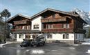 A typical alpine house with a wooden facade and balconies. In front of the house are several cars, surrounded by snow-covered ground.