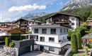 Ein modernes Haus mit zwei Etagen in einer malerischen Berglandschaft. Im Hintergrund sind hohe Berge und weitere Wohnhäuser sichtbar.