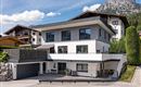 Ein modernes Haus mit großen Fenstern und einem klaren Parkplatz. Im Hintergrund sind Berge und eine schöne Landschaft sichtbar.