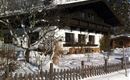 Ein traditionelles Holzhaus im Schnee mit einem grünen Baum im Vordergrund. Die Landschaft ist klar und sonnig, mit Bergen im Hintergrund.