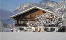 A traditional wooden house in winter, surrounded by snow. Mountains can be seen in the background.