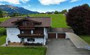 A charming alpine house with a large garden and plenty of green space. In the background, gentle hills and a blue sky can be seen.