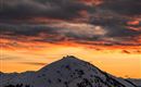 Ein schneebedeckter Berg unter einem orangefarbenen Sonnenuntergang. Die Wolken sind dramatisch und verleihen der Szene eine mystische Stimmung.